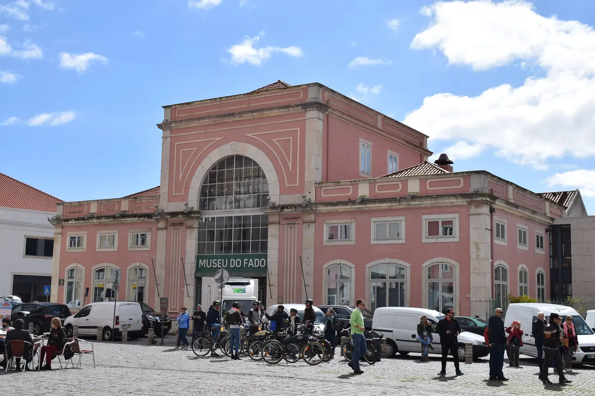Edificio neoclásico rosa del Museu do Fado al borde de una plaza empedrada, con personas y furgonetas aparcadas delante y un cielo azul al fondo